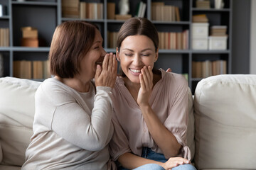 Middle-aged mother telling on ear to adult daughter secret, different generation women sit on sofa in living room share private hidden info laughing feels overjoyed, funny spend time together at home