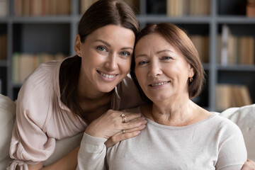 Close up different age generations women, adult daughter hugs from behind seated on sofa elderly mother, family portrait, photoshoot indoors, relatives closest people, resemblance and heredity concept - obrazy, fototapety, plakaty