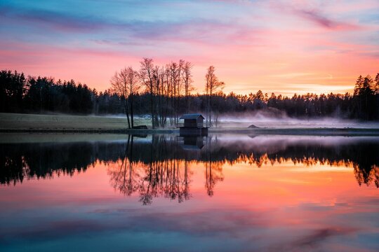 a lake at sunset reflecting a forest surrounding it with mist
