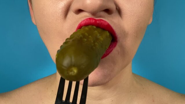 Unrecognizable young woman vibrantly colored lips enjoys a crunchy bite of a pickled cucumber, savoring tangy flavor indoors. Close-up mouth red lipstick savoring pickle isolated on blue background