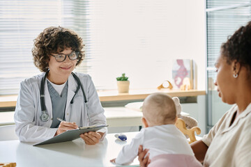 Obraz premium Professional pediatrician writing prescription while mother sitting with baby at the table