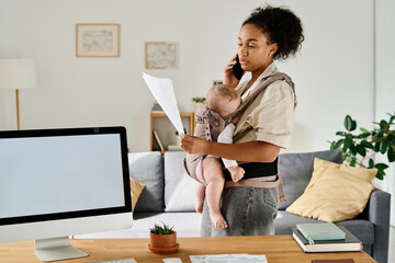 Young mom talking on the phone and reading document, she working at home while sitting with baby - obrazy, fototapety, plakaty