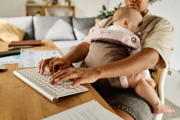 Young woman typing on keyboard while her baby sleeping in sling, she working at home during...