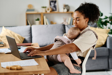 Young mother sitting at table and working online on laptop at home while her baby sleeping in sling