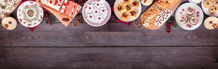 Christmas food top border. Above view on a dark wood banner background. Assortment of appetizers and delicious sweets. Copy space. Holiday party food concept.