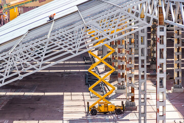 Workers install the metal sheet on the trusses and the metal frame of the warehouse roof. Metal frame high rise construction with diesel scissor lift - obrazy, fototapety, plakaty