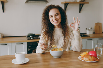 Young woman of African American ethnicity wear casual clothes sweater eat breakfast muesli cereals with milk fruit in bowl show ok sit at table in kitchen at home alone Lifestyle cooking food concept