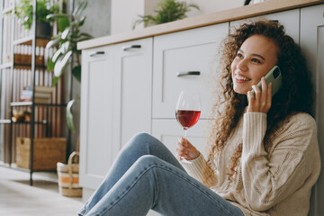 Close up young housewife woman of African American ethnicity wear casual clothes sweater drink wine talk on mobile cell phone sit near table in kitchen at home alone. Lifestyle cooking food concept.