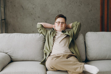 Young smiling man with down syndrome wear glasses casual clothes hold hands behind neck sit on grey...
