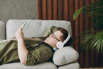 Young man with down syndrome wear clothes headphones listen music use mobile cell phone lay on grey sofa couch stay at home flat rest spend free time in living room. Genetic disease world day concept.