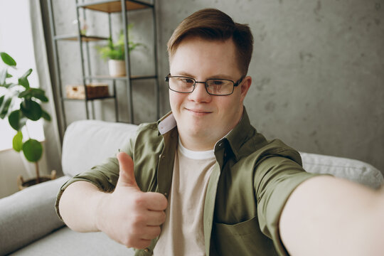 Young man with down syndrome wears casual clothes do selfie shot on mobile cell phone sits on grey sofa couch stay at home flat rest spend free time in living room. Genetic disease world day concept.