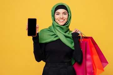 Young happy arab muslim woman wear green hijab abaya clothes hold shopping package bag blank screen mobile cell phone isolated on plain yellow background. Black Friday sale buy day, uae islam concept