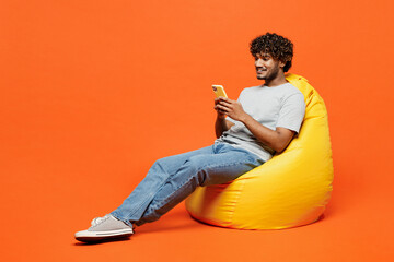 Full body young smiling happy Indian man he wears t-shirt casual clothes sit in bag chair use mobile cell phone type message isolated on orange red color background studio portrait. Lifestyle concept.