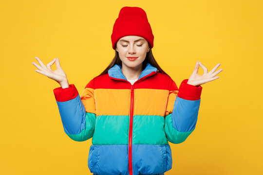 Young spiritual woman wear padded windbreaker jacket red hat casual clothes hold spread hands in yoga om aum gesture relax meditate try calm down isolated on plain yellow background Lifestyle concept