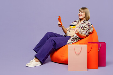 Full body elderly woman in beige shirt casual clothes sit in bag chair hold mobile phone credit card shopping online package bag isolated on plain purple background Black Friday sale buy day concept