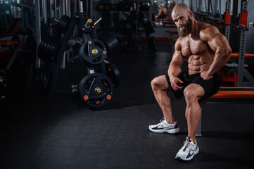 A muscular bald man in shorts is resting on a bench after a workout. Bodybuilder showing off his...