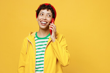 Young happy woman wears waterproof raincoat outerwear red hat talk speak on mobile cell phone isolated on plain yellow background studio portrait. Outdoors lifestyle wet fall weather season concept.