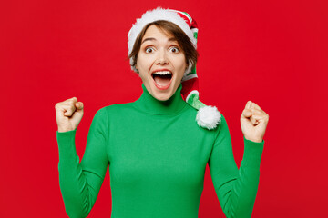 Young woman wear warm cozy green turtleneck Santa hat posing do winner gesture celebrate clench fist say yes isolated on plain red background Happy New Year 2024 celebration Christmas holiday concept