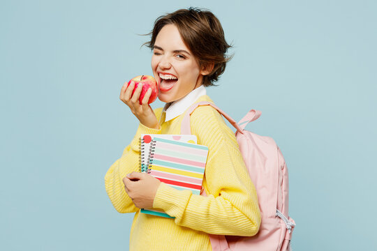 Side view young woman student wear casual clothes yellow sweater backpack bag hold books notebooks eat apple wink blink eye isolated on plain blue background. High school university college concept