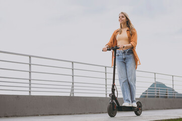 Full body young happy confident calm fun woman wearing orange shirt casual clothes riding e-scooter...