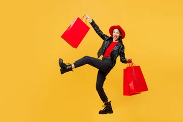 Fotobehang Full body side view young happy smiling woman wear casual clothes red hat hold shopping paper package bags walk go raise up hand isolated on plain yellow background. Black Friday sale buy day concept. © ViDi Studio