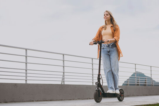 Full body young happy confident calm fun woman wearing orange shirt casual clothes riding e-scooter transport walking rest relax in spring green city park outdoors on nature. Urban lifestyle concept.