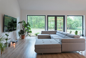 Comfortable sofa, tv on the wall, wooden floor and large window in modern interior of living room...