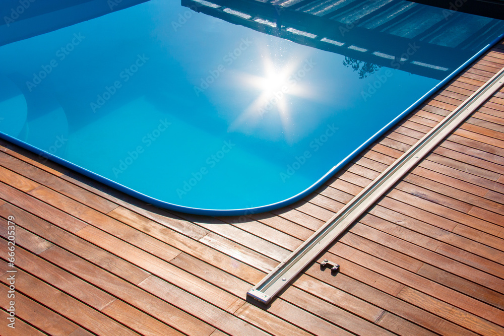 Wall mural ipe wood decking around the pool, edge of the outdoor swimming pool with sun reflection on the blue 