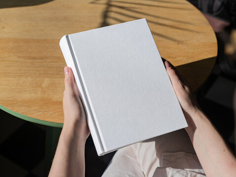 Book cover mockup, hand holding blank clean hardcover mock up on wood desk