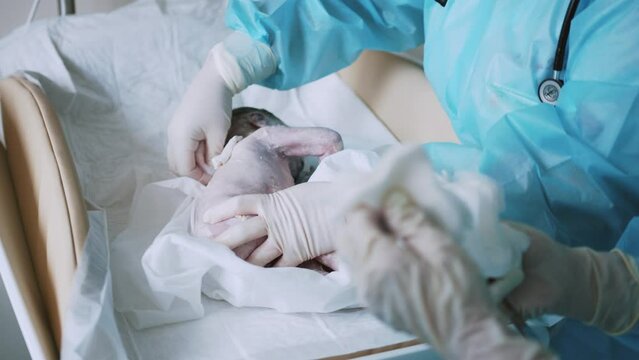 Newborn cute infant baby with hand holding clip on umbilical cord at a hospital nursery. Nurse on gloves cut and tie up umbilical cord to newborn crying baby in maternity hospital after birth
