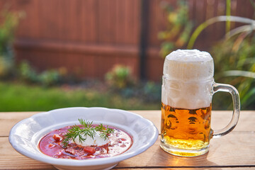 Russian or ukrainian borsch soup served outside with mug of pilsner beer in the garden restaurant in summer sunny day. Beetrot soup with sour cream is traditional recipe from east eauropean cuisine.