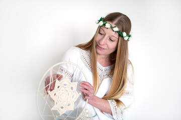 The innocent image of a young woman with long hair in a pink wreath and a white dress on a white background in the studio, in the hands of a dreamcatcher of pastel cotton threads. Purity and Harmony.