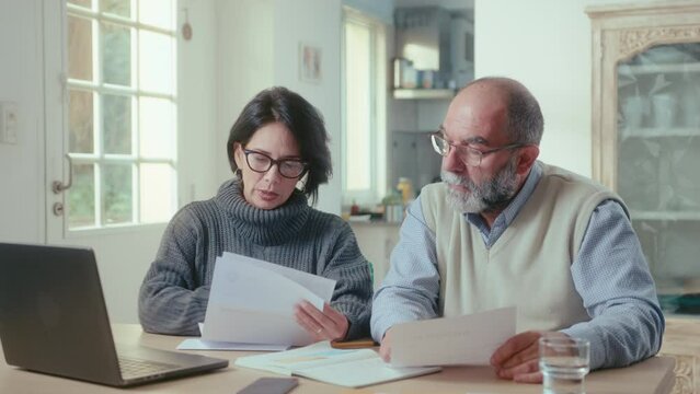 Senior couple sitting at table with laptop, reading papers and discussing family budget plan during the day at home. Medium shot