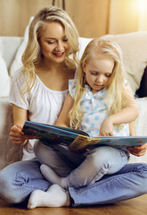 Happy family. Blonde young mother reading a book to her cute daughter while sitting at wooden floor in sunny room. Motherhood concept