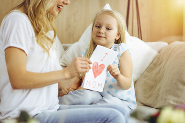 Happy mother day in sunny flat. Child daughter congratulates mom and gives her postcard with heart drawing. Family concept