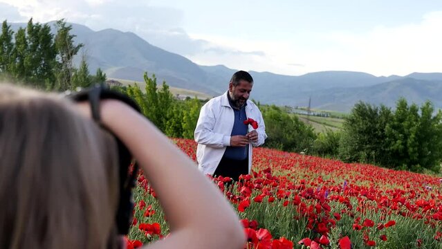 A young girl photographs an adult Middle Eastern man with a beard in a field of red poppies in the mountains. Photo shoot of a cute adult man picking red flowers and smiling.
