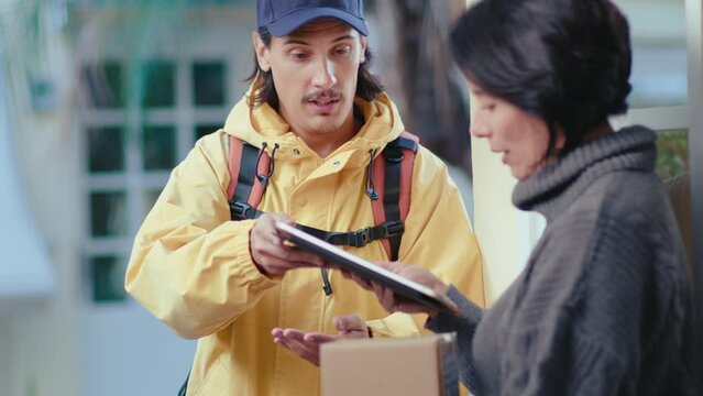 Male courier giving parcel to mature woman and asking her to put a signature on a digital tablet to verify received order. Medium shot, rack focus