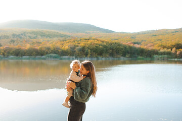 Happy stylish woman holding newborn baby over lake outdoor in sun light. Motherhood. Autumn season.