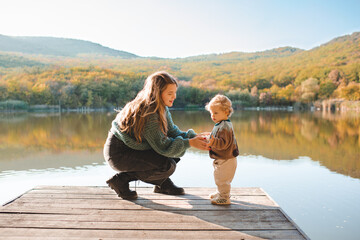 Happy stylish woman holding newborn baby over lake outdoor in sun light. Motherhood. Autumn season.