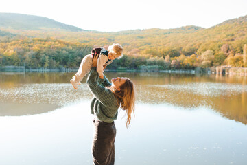 Happy stylish woman holding newborn baby over lake outdoor in sun light. Motherhood. Autumn season.