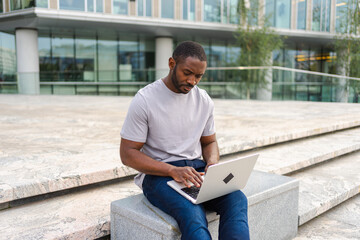 African American man freelancer using laptop typing chatting on urban street in city. Guy having virtual meeting online chat conference. Entrepreneur programmer working on laptop outdoors. Remote work