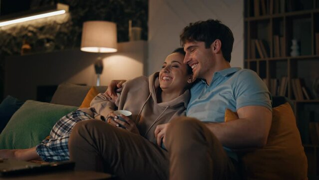 Smiling spouses watching tv sitting at cozy living room with coffee late evening