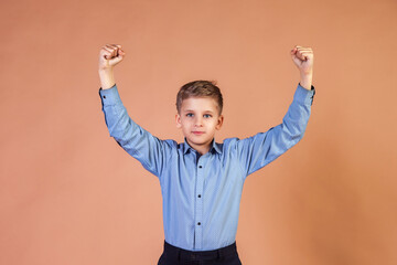 Winner school boy 10 year old in blue shirt arms raised at beige background, looking at camera. Guy...