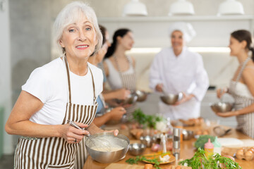 Elderly lady holding bowl and whisk in her hands surrounded by other female members during cooking master class