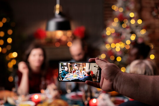 Person takes photos of people at table enjoying dinner festivity with glasses of alcohol surrounded by xmas tree and ornaments. Diverse friends and family posing for pictures on christmas eve.