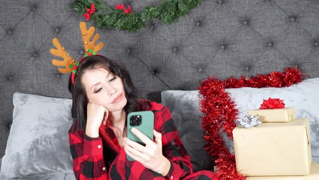 Young beautiful woman in red plaid pyjama wearing antlers headband, scrolling on phone borred in Christmas decorated room with presents.