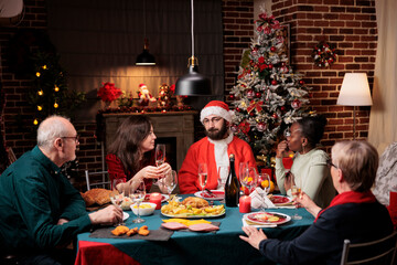 Person santa claus cosplay at dinner making people laugh and celebrating christmas eve festivity together. Senior people, friends and family gathering around table to eat and drink on holiday.