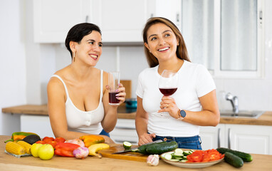 Obraz na płótnie Canvas Smiling young Hispanic woman cooking with sister in home kitchen, chatting cheerfully and drinking wine while preparing vegetable salad. Happy family relationship concept..