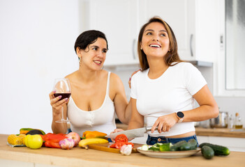 Obraz na płótnie Canvas Happy carefree young latin american lesbian couple chatting carelessly and drinking wine while preparing vegetable salad for dinner in cozy kitchen interior at home