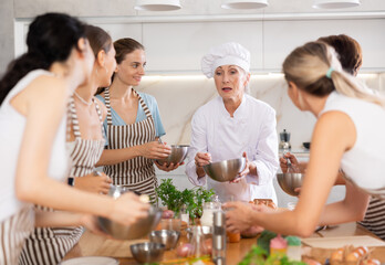 Obraz na płótnie Canvas Elderly woman presenter of cooking courses for amateurs holds bowl in hands and explains to female participants principles of proper mixing 